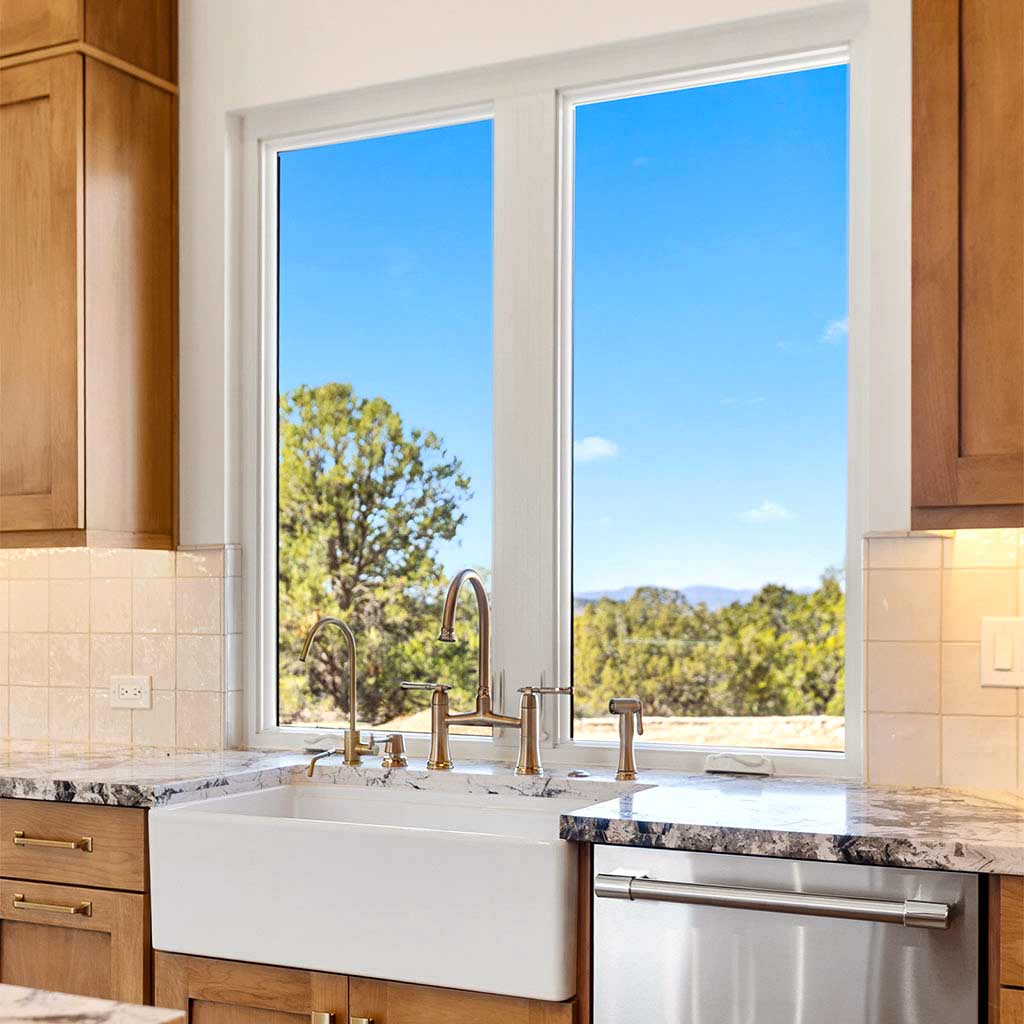 White apron-front sink under a large window in a bright kitchen, with brass faucet fixtures and marble countertops.