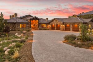 Luxurious stone-and-wood mansion with a curved driveway leading to multiple wooden garage doors at sunset.